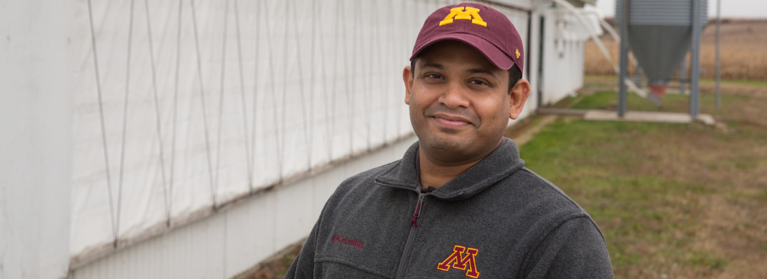 A man wearing a UMN hat and sweatshirt stands in front of a white building and grass smiling